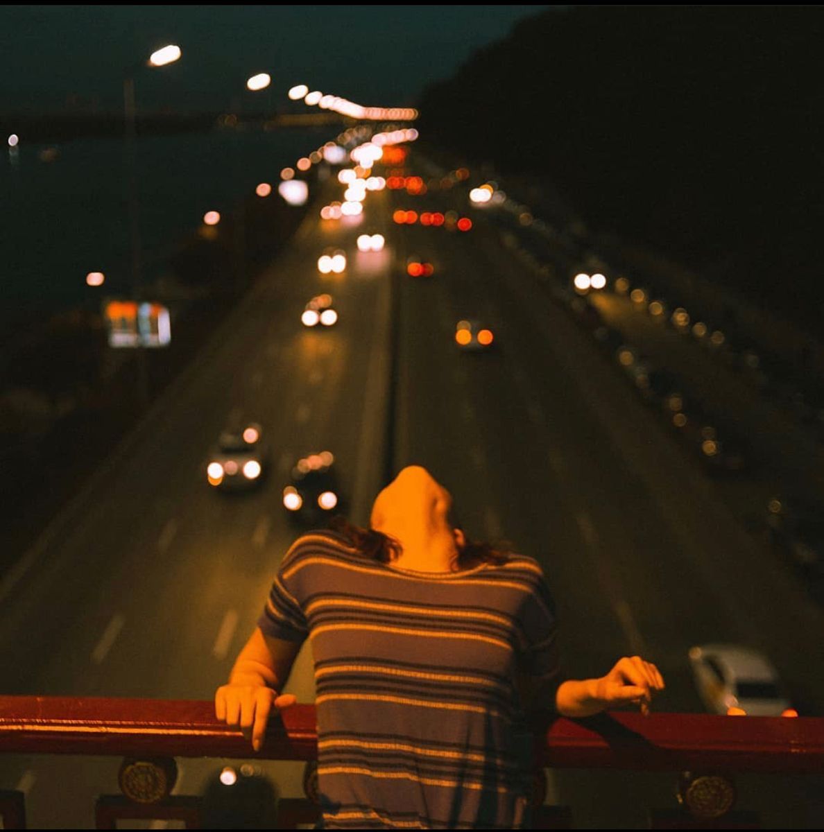A young person leans back over a freeway overpass at night, headlights blurring behind them.